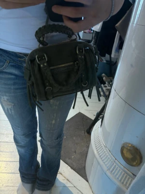 Svart minihandväska med flätat handtag - Säljer en svart minihandväska i mocka-look med flätat handtag och justerbar axelrem. Väskan har silverfärgade metalldetaljer, dragkedja framtill och dekorativa remmar. Perfekt för dig som vill ha en liten men cool accessoar./köptes i London 