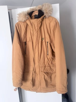 Beige parkas från Lyle & Scott och mössa  - Snygg beige parkas från Lyle & Scott med avtagbar huva och fuskpäls. Jackan har två bröstfickor med dragkedja, två stora framfickor och gul logga på bröstet. Perfekt för kalla dagar med sin vadderade insida och klassiska design. Xxl och mössa på köpet