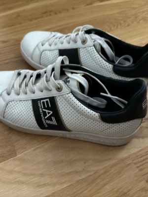 Vita EA7 Emporio Armani sneakers - Snygga vita sneakers från EA7 Emporio Armani med svart detalj och logga på sidan. Skorna har perforerat läder för extra ventilation, vita snören och svart hälparti. Perfekt för dig som vill ha en clean och stilren look.
