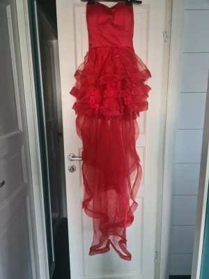 Röd balklänning med volanger - Snygg röd långklänning med volanger och transparent, asymmetrisk kjol. Klänningen är axelbandslös och har snörning i ryggen för en justerbar passform. Perfekt för dig som vill sticka ut med en dramatisk look. Har en liten repa i botten av klänningen som man kan se på sista bilden. Köpte den begagnad men har kvar prislappen från erikshjälpen är kvar, aldrig använd av mig.