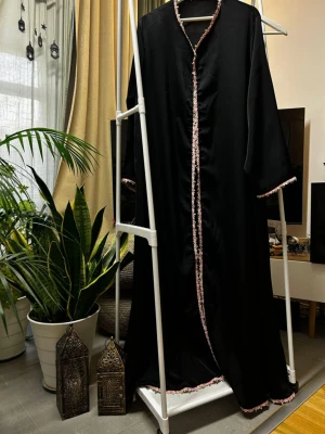Svart satin abaya & sjal - Elegant svart satinabaya med vackra rosa broderade detaljer längs kanterna.  Tillverkad i mjukt och följsamt satintyg som ger ett lyxigt och stilrent intryck. Modellen är lång, öppen framtill.