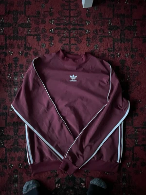 Vinröd Adidas sweatshirt med vita ränder - Säljer en vinröd Adidas sweatshirt med klassiska vita ränder längs ärmarna och ryggen. M storlek