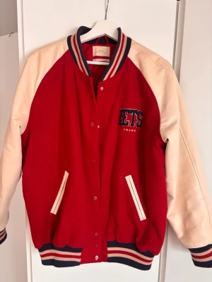 Röd och beige bomberjacka LETS - Snygg röd bomberjacka från Urban outfitters med beigea ärmar i skinnimitation och broderad text på bröstet. Jackan har randiga muddar i rött, beige och marinblått samt två fickor framtill. Stängs med tryckknappar och har en klassisk college-look. Den är overaized så passar många och har ull i sig + är fodrad, så den e varm🙌🫶