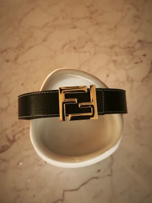 Svart skärp från Fendi med guld märke - Snyggt svart skärp från Fendi med en stor, ikonisk guldfärgad Fendi-logga som spänne. Skärpet har vita sömmar längs kanterna för en extra stilren look. Perfekt accessoar för att lyfta vilken outfit som helst.