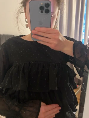 Svart söt glittrig volangblus! 💘 - Säljer en svart blus med transparenta långärmar och små prickar i tyget. Blusen har flera volanger framtill och är gjord i ett lätt, skirt material som ger en luftig känsla! Storlek xxs och sälj inte längre! Jätte söt ! ❤️