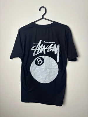 Svart Stussy t-shirt med 8-ball print - Svart t-shirt från Stussy med stort vitt 8-ball tryck och logga på ryggen samt mindre tryck på bröstet. Klassisk rund hals och korta ärmar. Tillverkad i mjuk bomull som känns skön mot huden. Perfekt för dig som gillar streetwear och vill sticka ut.