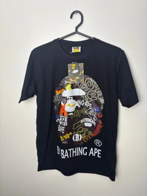 Svart BAPE t-shirt med grafiskt tryck - Cool svart t-shirt från BAPE med stort grafiskt tryck på bröstet i flera färger och olika mönster. Klassisk rund hals och korta ärmar. Materialet är mjuk bomull och logotypen syns tydligt både på trycket och etiketten.