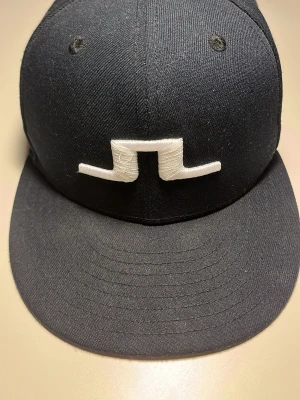 Mörkblå J.Lindeberg x New Era keps limited edition.  - Säljer en svart fitted keps från J.Lindeberg i samarbete med New Era 59FIFTY. Kepsen har ett vitt broderat J.Lindeberg-logo framtill och diskret broderad text bak. Klassisk rak skärm och snygg minimalistisk look. Kepsen är limited edition därav priset.