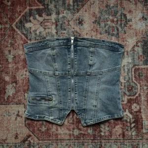 Jeans korsett liknande topp med dragkedja och sömn detaljer  - Säljer en snygg korsettopp i blå denim med markerade sömmar och dragkedja framtill. Toppen har en  liten ficka och är axelbandslös.
