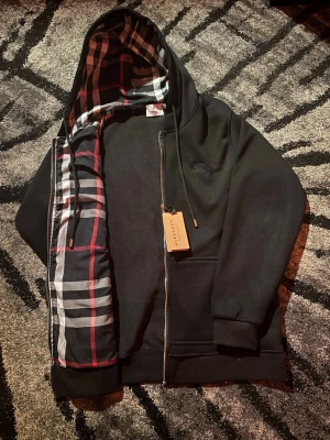 Svart hoodie med rutigt foder Burberry - Svart hoodie från Burberry med dragkedja och klassiskt rutigt foder i huvan och längs insidan. Diskret Burberry-logga på bröstet, ribbade muddar och fickor framtill. Snygg och tidlös design med exklusiv känsla.
