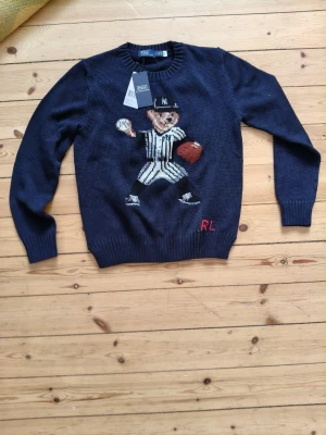 Mörkblå stickad tröja Polo Ralph Lauren - Mörkblå stickad tröja från Polo Ralph Lauren med en stor broderad björn i basebollkläder på framsidan. Tröjan har ribbade muddar vid ärmslut och nederkant samt RL-broderi i rött. Rund halsringning och klassisk passform.