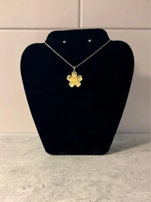 Silverhalsband med gul blomma - Snyggt halsband med en silverkedja och ett hängsmycke i form av en gul blomma. Blomman har fem kronblad och en liten detalj i mitten. Perfekt för dig som vill ha en söt och unik accessoar till din outfit. 