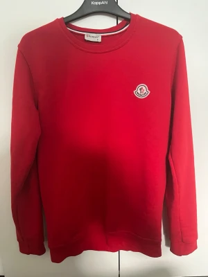 Röd sweatshirt från Moncler - Snygg röd sweatshirt från Moncler med rund halsringning och Moncler-logga på bröstet. Tröjan har långa ärmar och ribbade muddar vid ärmslut och nederkant. Perfekt för en avslappnad och trendig stil.