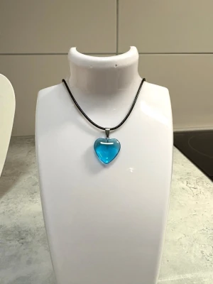Halsband med blått hjärthänge - Snyggt halsband med ett genomskinligt blått hjärta som hänge. Hjärtat är gjort av glasliknande material och hänger i en tunn svart rem. Perfekt accessoar för att ge din outfit en färgklick och en söt touch. Hänge 20mm. 