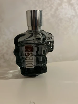 Diesel Only The Brave parfym - Cool parfym från Diesel i en unik flaska formad som en knytnäve. Flaskan är mörkgrå i glas med en silverfärgad metallkork och Diesel-loggan i metall på sidan. Perfekt för dig som gillar statement-detaljer på hyllan.