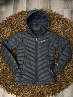 Svart dunjacka Peak Performance dam - Snygg svart dunjacka från Peak Performance med huva och dragkedja framtill. Jackan har två sidofickor med dragkedja och logga på bröstet. Lättviktigt material och quiltad design gör den perfekt för kalla vinterdagar.