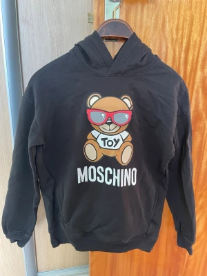 Svart Moschino hoodie med björntryck - Svart hoodie från Moschino med en stor björn med röda solglasögon och texten 'TOY' på magen. MOSCHINO-logga i vitt under motivet. Klassisk huva och ribbade muddar. Perfekt statement-plagg för dig som gillar lekfull design.Passar runt 12-13 åringar,som är runt 135-150 cm skulle jag säga så ganska liten. Fint skick inga fläckar eller så. 