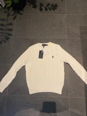 Vit kabelstickad tröja Polo Ralph Lauren - Kabelstickad vit tröja från Polo Ralph Lauren med rund halsringning och den klassiska broderade loggan på bröstet. Tröjan har ribbade muddar vid ärmslut och nederkant, vilket ger en snygg och tidlös look. Perfekt att matcha med jeans eller kjol.