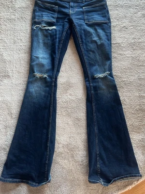 Blå utsvängda jeans Gina Tricot - Säljer ett par blå lågmidjade bootcut jeans från Gina Tricot med slitna detaljer på låren och små fickor fram och bak. Jeansen är väldigt låga och nästintill oanvända,  använt de ungefär 4-5 gånger. Perfekta för dig som gillar Stockholmsstil och lågmidjat. 