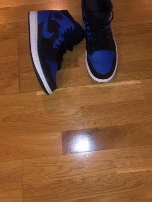 Air Jordan 1 Mid Svart/Blå - Säljer ett par Air Jordan 1 Mid i svart och blått med vit sula. Klassisk siluett med svart läder och blå detaljer, inklusive swoosh och tåbox. Skorna har snörning och ikoniska Jordan-loggor på sidan och plösen. Perfekta för dig som gillar streetwear och sneakerskultur.