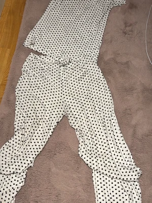 Vit prickig pyjamas med kort ärm - Supersöt pyjamas i vitt med svarta prickar. Setet består av en kortärmad topp och långa byxor med resår i midjan. Materialet känns mjukt och skönt, perfekt för mysiga kvällar hemmae. Klassisk och lekfull design som passar dig som gillar retrostil.