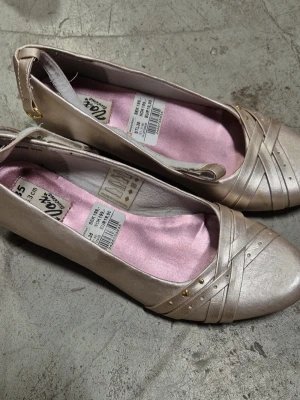 Ballerinaskor - Säljer ett par champagnefärgade ballerinaskor från Skopunkten med låg kilklack. Skorna har dekorativa band och små guldfärgade nitar framtill samt en mjuk rosa innersula. Perfekta för dig som vill ha något stilrent men ändå lite extra.