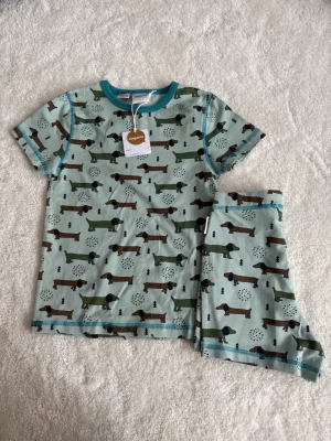 Grönblå pyjamas med taxar, Maxomorra - Kortärmad pyjamas från Maxomorra i mjuk ekologisk bomull. Setet är ljusgrönt med ett gulligt taxmönster i grönt och brunt samt turkosa detaljer vid hals och benslut. Perfekt för mysiga kvällar och sköna nätter.