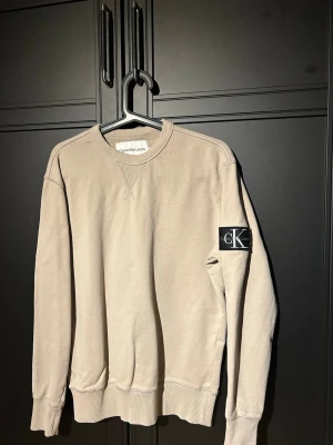 Beige sweatshirt från Calvin Klein - Snygg beige sweatshirt från Calvin Klein Jeans med rund halsringning och ribbade muddar. Tröjan har en svart patch med CK-logga på ärmen och är tillverkad i mjuk bomull. Perfekt för en avslappnad och stilren look. Storlek S och i bra skick 7/10