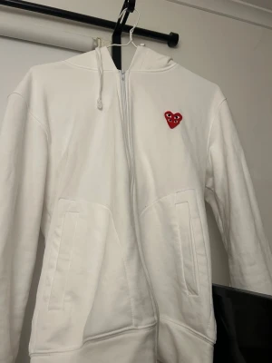 Vit hoodie Comme des Garçons Play - Säljer en vit hoodie från Comme des Garçons Play med dragkedja, huva och två fickor framtill. På bröstet sitter det klassiska röda hjärtat med ögon. Hoodien är i mjukt bomullsmaterial och har en clean, stilren look som passar perfekt till streetwear.
