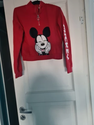 Röd Mickey Mouse hoodie med dragkedja - Cool röd hoodie från Disney med stor Mickey Mouse-tryck på framsidan och texten 'MICKEY' i vitt längs ena ärmen. Tröjan har halv dragkedja med ringdetalj och ribbade muddar. Perfekt för dig som gillar retro och streetstyle.