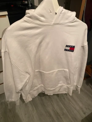 Vit hoodie från Tommy Jeans - Snygg vit hoodie från Tommy Jeans med klassisk logga på bröstet i blått, vitt och rött. Hoodien har stor huva, magficka och ribbade muddar vid ärmar och nederkant. Perfekt för en avslappnad och stilren look.