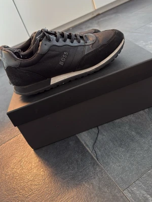 Svarta sneakers från BOSS - Snygga svarta sneakers från BOSS med klassisk låg profil. Skorna har en mix av mocka och textil på ovandelen, svarta skosnören och vit sula med svart yttersula. Perfekta för en stilren och modern look.