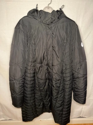 Svart quiltad kappa med huva - Lång svart quiltad kappa från Outdoor med huva och dragkedja framtill. Kappan har ett stilrent mönster och en patch på ärmen. Perfekt för kyliga dagar och enkel att matcha med olika outfits.