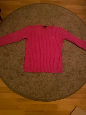 Rosa kabelstickad tröja Ralph Lauren - Säljer en rosa kabelstickad tröja från Ralph Lauren med gul broderad RL-logga på bröstet. Tröjan har rund halsringning, långa ärmar och ribbade muddar. Perfekt för dig som gillar klassisk färgglad stil.