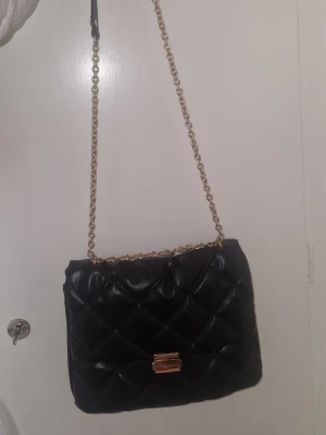 Axelväska  - Snygg svart axelväska i quiltat mönster med guldfärgad kedjerem och matchande guldfärgat spänne. Väskan har en klassisk rektangulär form och är tillverkad i ett glansigt material som liknar konstläder. Perfekt för dig som gillar stilrena accessoarer.
