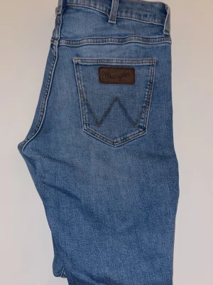 Blå jeans från Wrangler - Klassiska blå jeans från Wrangler i storlek W30/L32 med rak passform och fem fickor. Jeansen har en snygg tvättad look och detaljerad bakficka med Wrangler-logga i läder. Tillverkade i slitstarkt denimtyg, perfekta för en avslappnad och trendig stil.