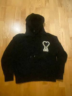 Svart Ami Paris hoodie med hjärtdetalj - Säljer en svart hoodie från Ami Paris med stor vit broderad hjärtdetalj och bokstaven A på bröstet. Hoodien har dragsko i huvan och ribbade muddar vid ärmar och nederkant. Perfekt för dig som gillar streetwear och vill ha något stilrent men ändå unikt. Nästan helt oanvänd. 