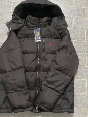 Svart pufferjacka från Polo Ralph Lauren - Säljer en svart pufferjacka från Polo Ralph Lauren med huva och flera dragkedjeförsedda fickor. Jackan har det klassiska röda Polo-logot på bröstet och quiltad design. Perfekt för kalla dagar och riktigt snygg till streetwear-stilen.