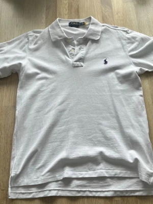 Vit piké från Polo Ralph Lauren - Klassisk vit pikétröja från Polo Ralph Lauren med korta ärmar, krage och knappar framtill. Ikoniska blå loggan broderad på bröstet. Tillverkad i mjuk bomull för en bekväm känsla och clean look.