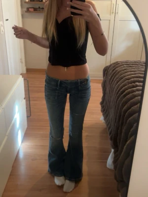 Blå bootcut jeans med låg midja - Säljer ett par blå bootcut jeans med låg midja och lätt slitna detaljer på låren. Jeansen har klassisk femficksdesign och är tillverkade i denim med lite stretch för skön passform. Perfekta för en avslappnad och trendig look.