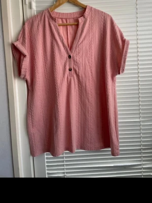 Rosa kortärmad blus med strukturmönster - Söt rosa blus med korta ärmar och dekorativa knappar framtill. Blusen har ett subtilt strukturmönster i tyget och en avslappnad passform med v-ringad hals. Perfekt för en chill och trendig look.