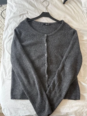 Mörkgrå kofta med knappar  - Mysig mörkgrå kofta  med rund halsringning och knäppning framtill. Koftan har långa ärmar och är tillverkad i ett mjukt, stickat material som passar perfekt för lager på lager. Enkel och stilren design som funkar till det mesta.