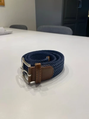 Marinblått flätat bälte från VB - Snyggt marinblått flätat bälte från VB med detaljer i brunt skinn och silverfärgat spänne. Perfekt accessoar för att lyfta din outfit med en stilren touch. Bältet har en flexibel design och klassisk look.