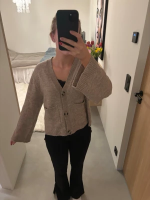 Beige kofta med fickor och knappar - Beige kofta med v-ringning och stora knappar framtill. Koftan har två fickor och är stickad i ett mjukt material. Ärmarna är vida🫶🏻