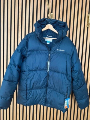 Mörkblå pufferjacka från Columbia - Snygg mörkblå pufferjacka från Columbia med huva och dragkedja framtill. Jackan har en quiltad design och är tillverkad i ett slitstarkt, vattenavvisande material som håller dig varm under kalla dagar. Perfekt för dig som vill ha en stilren och funktionell vinterjacka. Köpt till min sambo som julklapp men den var för stor och glömde skicka tillbaks den. Nypris 1900kr. Kom med bud. 
