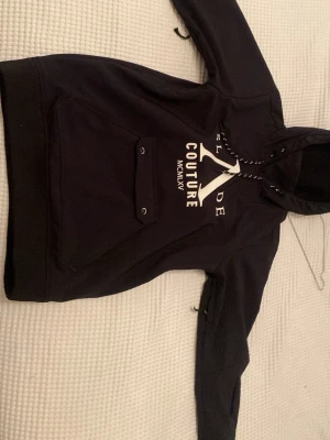 Svart skidjacka från Slade Couture - Svart skidjacka från Slade Couture storlek 36/S med stor vit logga och text på både fram- och baksidan. Tröjan har huva med dragsko och en praktisk magficka med knappdetaljer. Tillverkad i mjukt material som känns skönt mot huden. Perfekt för en avslappnad och trendig stil.