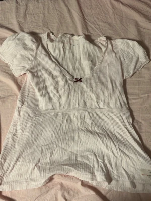 Vit ribbad topp med rosett och tryck - Söt vit ribbad topp med v-ringning och liten röd rosett framtill. Korta ärmar och ett tryck med texten 'Lost on Purpose' och blå detaljer på sidan. Perfekt för en chill och avslappnad stil.