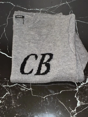 Cole Buxton tröja - Säljer en grå stickad tröja från cole Buxton med svarta bokstäver 'CB' på bröstet. Tröjan har rund halsringning, långa ärmar och ribbade muddar vid ärmslut och nederkant. Perfekt för dig som gillar enkel men snygg design.