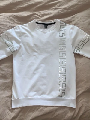 Vit tröja med silvriga detaljer - Vit sweatshirt med glansiga silvriga mönster längs ärmarna och en vertikal detalj framtill. Klassisk rund hals och ribbade muddar. PRISET KAN DISKUTERAS!!!!! Mvh 