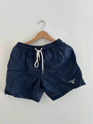 Marinblå badbyxor från Barbour - Snygga marinblå badbyxor från Barbour med vit dragsko i midjan och broderad logga på ena benet. Klassisk modell med elastisk midja och bakficka med lock. Perfekta för stranden eller poolen. Size S, knappt använda men legat i garderoben ett tag. Köpta i USA på Macy’s. 🚀🫧